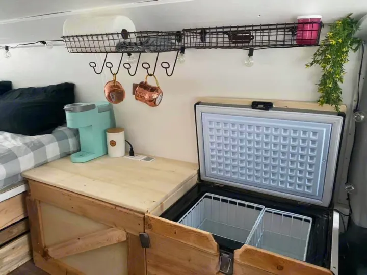 RV rental's compact kitchen area: light blue coffee maker, wood counter, open fridge with baskets, plus nearby bedding.. Ford Econoline 2012
