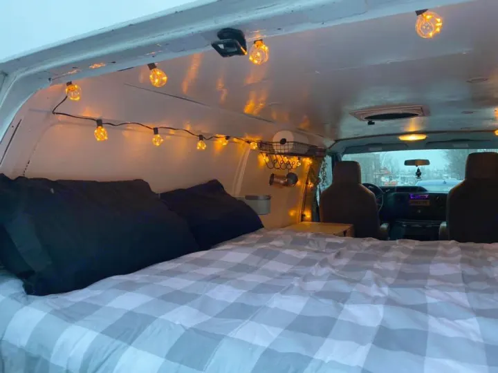 Step inside this cozy RV rental! A comfy bed with a checkered comforter, black pillows, & warm string lights create a welcoming camper van space.. Ford Econoline 2012