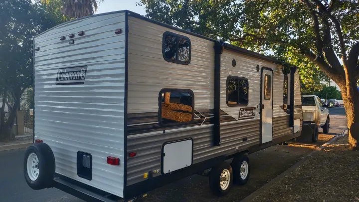 A light-colored Coleman travel trailer RV rental, equipped with dual axles and a rear spare tire, parked outdoors.. Dutchmen Coleman Lantern LT 2022