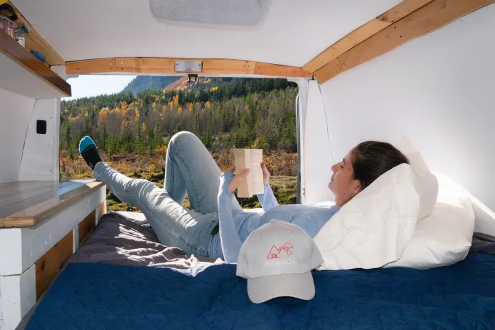 Enjoying an RV rental! A person reads in a cozy campervan bed, with fall forests and mountains visible outside.. Ford Econoline 2009