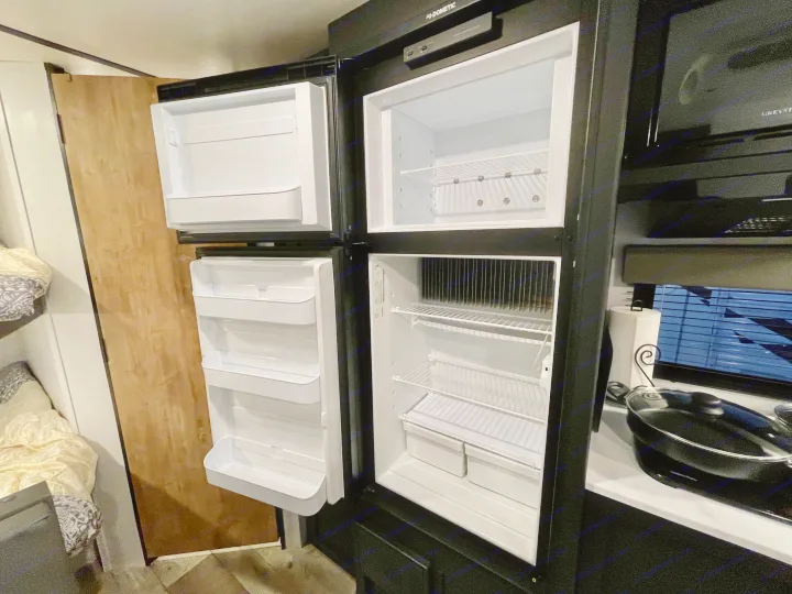 Open Dometic refrigerator inside an RV rental, revealing its empty shelves. Part of the compact kitchen and bunk beds are visible.. Keystone RV Springdale 2022