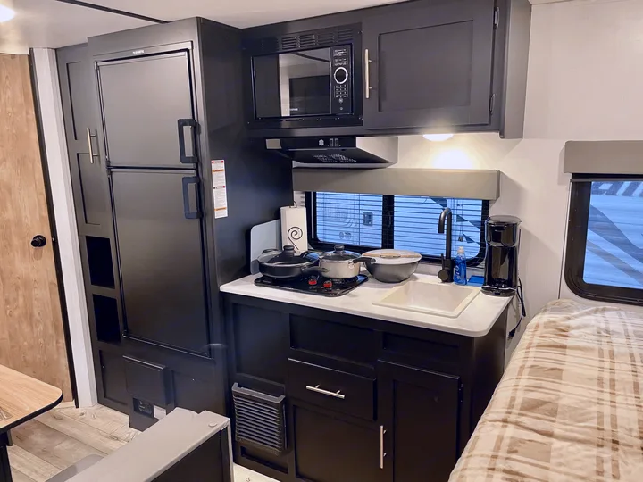 Explore this rv rental's compact kitchen with dark cabinets, fridge, microwave, and stove, next to a comfy bed.. Keystone RV Springdale 2022