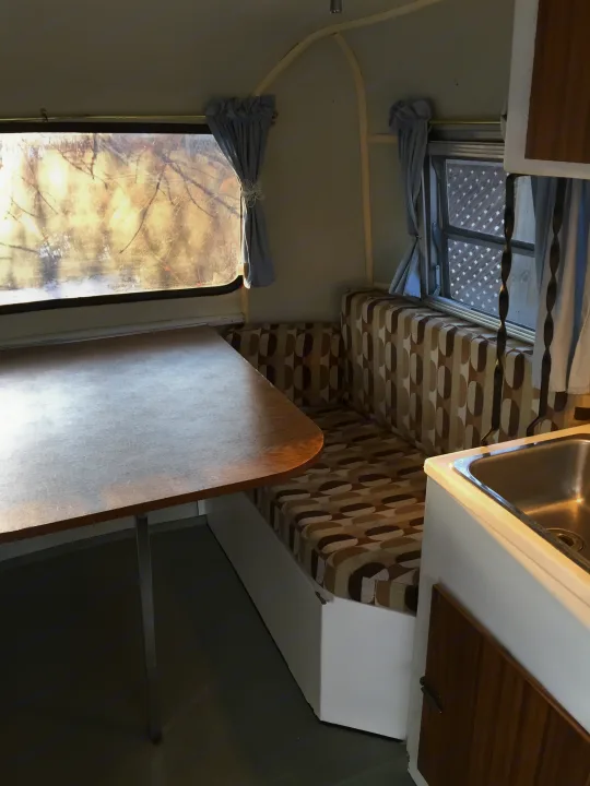 Cozy RV rental interior: a retro dining booth with patterned cushions, wooden table, and sink. Windows have light curtains.. Boler B13 1973