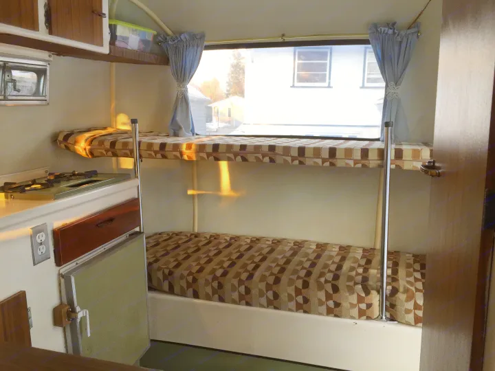 Interior view of an RV rental: two bunk beds with patterned cushions, a window with curtains, and a visible kitchen section.. Boler B13 1973