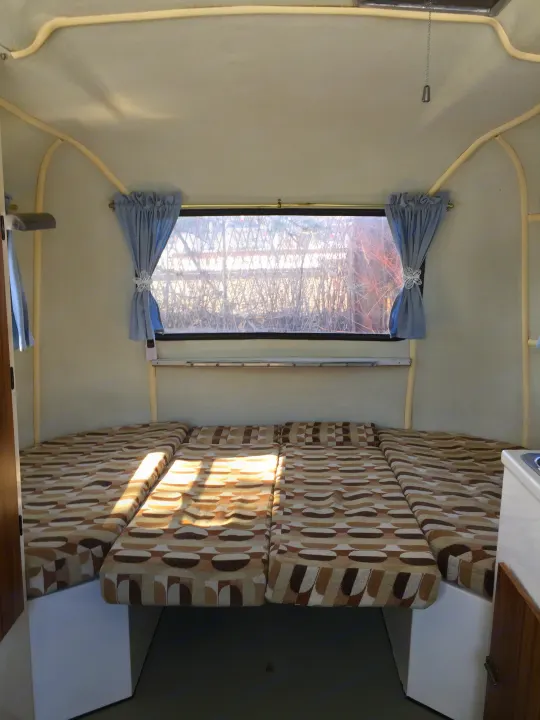 Bright RV rental interior featuring abstract patterned cushions, a window with blue curtains, and warm sunlight.. Boler B13 1973