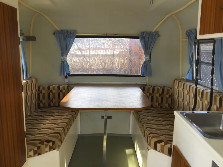 Inside a cozy RV rental: dining table, upholstered benches, sink, and windows with blue curtains.. Boler B13 1973