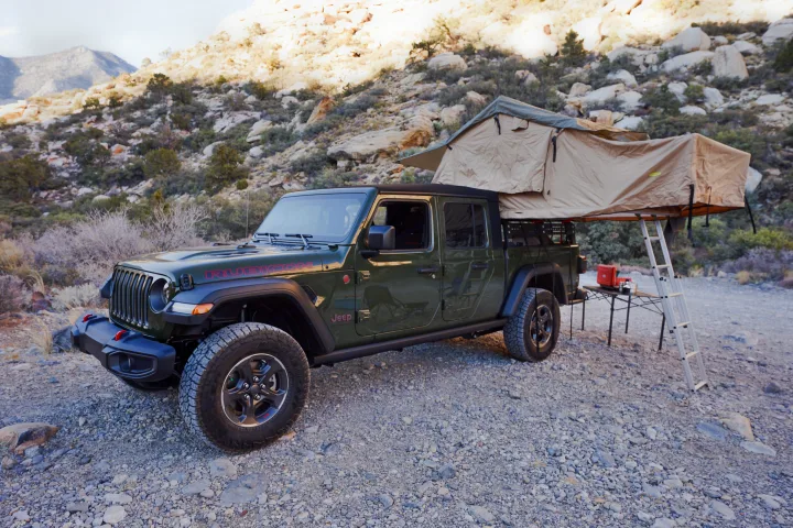 Green Jeep Gladiator with rooftop tent and camp setup on gravel, an ideal RV rental for off-road adventure camping.. Fiat Jeep Gladiator 2023