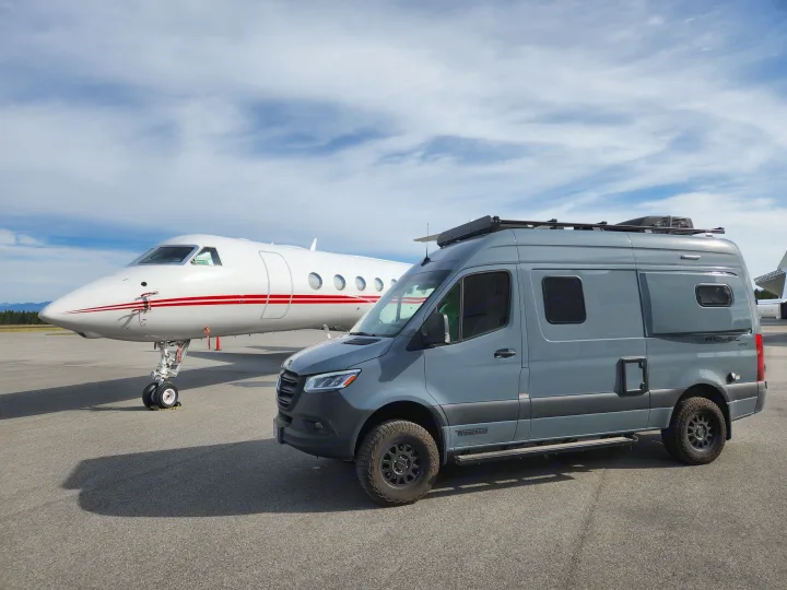 Experience adventure with an RV rental: a modern grey camper van parked next to a sleek white private jet on an airfield.. Winnebago Revel 2022