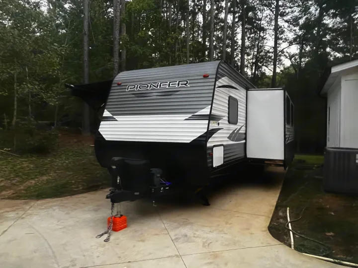 A modern grey and white Pioneer travel trailer, suitable for RV rental adventures, parked on concrete with trees. Awning extended, slide-out open.. Heartland RVs Pioneer 2020