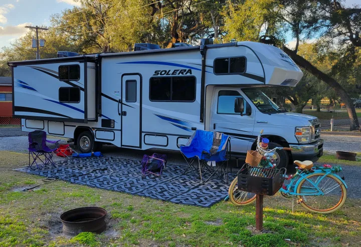 This rv rental is set up at a sunny campsite with chairs, a decorative bike, a grill, and a fire pit for outdoor relaxation.. Forest River Solera 2022