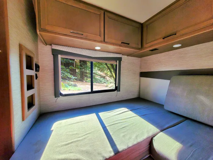 Bright RV rental interior: bed, window with forest view, overhead cabinets. Ideal for a serene travel getaway.. Thor Motor Coach Four Winds 2021