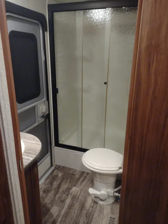 Compact RV rental bathroom with a frosted glass shower door, white toilet, and adjacent window view.. Heartland Trail Runner 2016