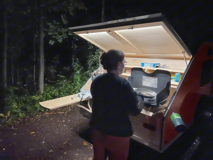 Night cooking at an RV rental campsite. A person uses a brightly lit, pull-out outdoor kitchen from a compact RV.. TC Tear Drop TC Original 2022