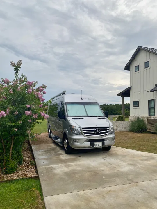 Silver Winnebago Sprinter RV rental parked on a Texas driveway next to a modern home and pink flowers.. Winnebago Era 2016