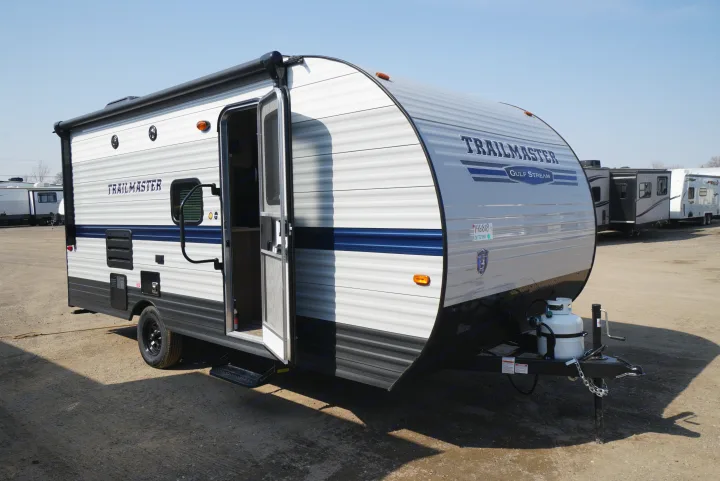 A Trailmaster Gulf Stream travel trailer (RV rental) is shown parked on a lot with its door open. Other RVs are visible in the background.. Gulf Stream Trailmaster 2022