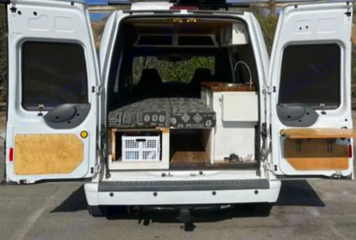 Rear of an open white camper van, showcasing a cozy bed, smart storage, and a sink setup. Ideal for an RV rental.. Ford Transit 2012