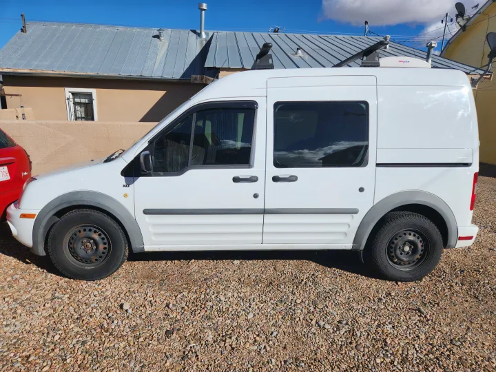 White Ford Transit Connect van with roof fan, possibly an rv rental, parked on gravel. A house with a metal roof is in the background.. Ford Transit 2012