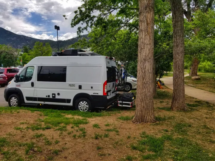 A white Ram 1500 camper van, an RV rental, parked amidst trees with mountains in the background, equipped with bikes and cargo on the back.. Thor Rize 2022