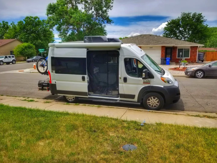 White Ram ProMaster RV rental, equipped with bike rack & open door screen, parked curbside. Perfect for your next adventure.. Thor Rize 2022