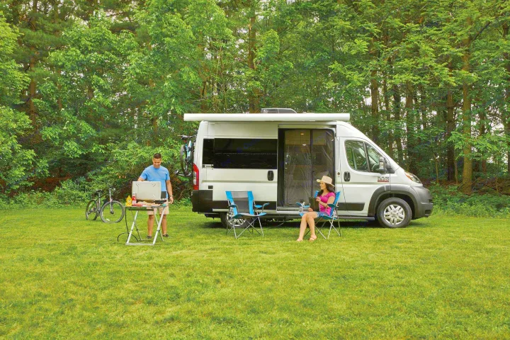 Couple enjoys outdoor grilling & relaxation with an RV rental at a scenic campsite. Perfect for nature lovers.. Thor Rize 2022