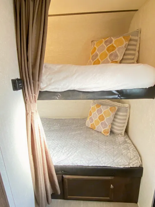Cozy RV rental bunk beds with colorful pillows, white bedding, and a privacy curtain. Features compact sleeping & lower storage.. Starcraft Launch 2018