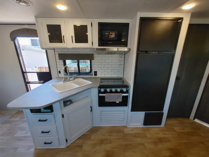 Modern RV rental kitchen interior: white cabinets, black appliances (fridge, stove, microwave), double sink, wood floor.. Keystone RV Hideout 2022