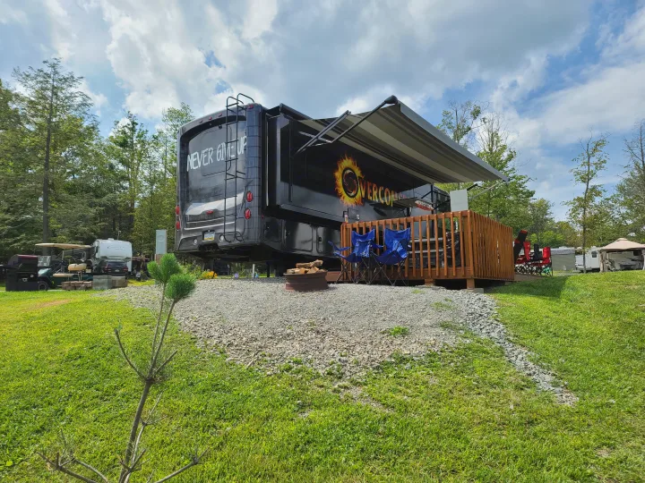 A large black RV rental with "OVERCOME" branding, an attached wooden deck, and fire pit on a gravel campsite with other campers.. Jayco Precept 2020