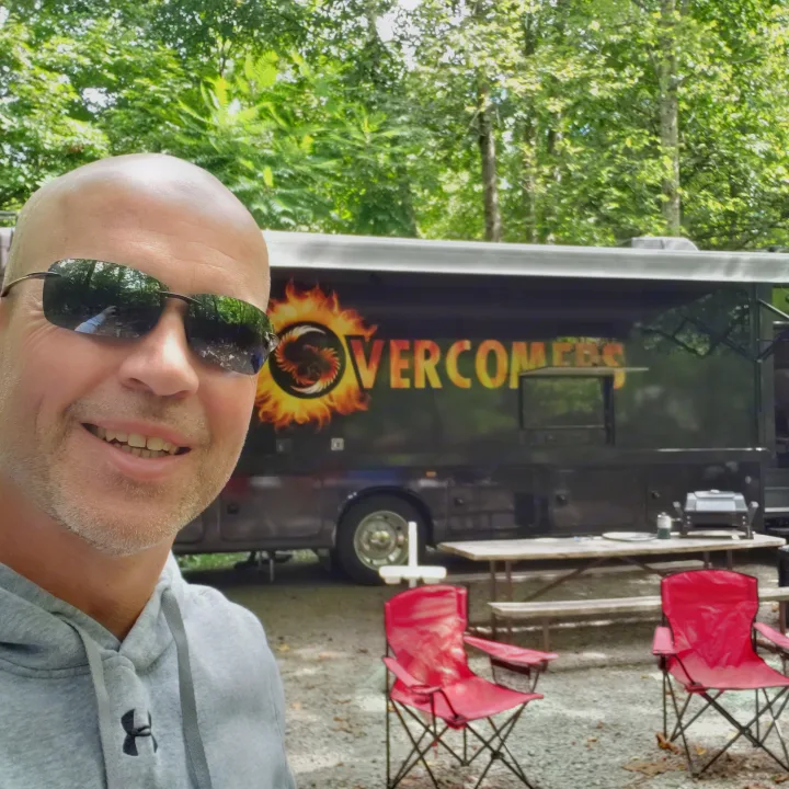 Man smiling in front of a black RV rental with "OVERCOMERS" logo, parked in a wooded area with picnic table and chairs.. Jayco Precept 2020