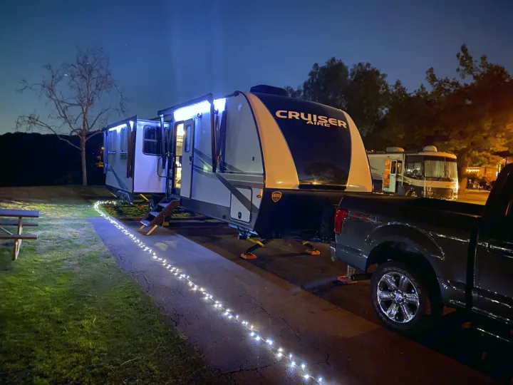 Night scene at an RV rental campsite: a lighted travel trailer with a pickup truck, a picnic table, and path lights.. Crossroads RV Cruiser Aire 2021