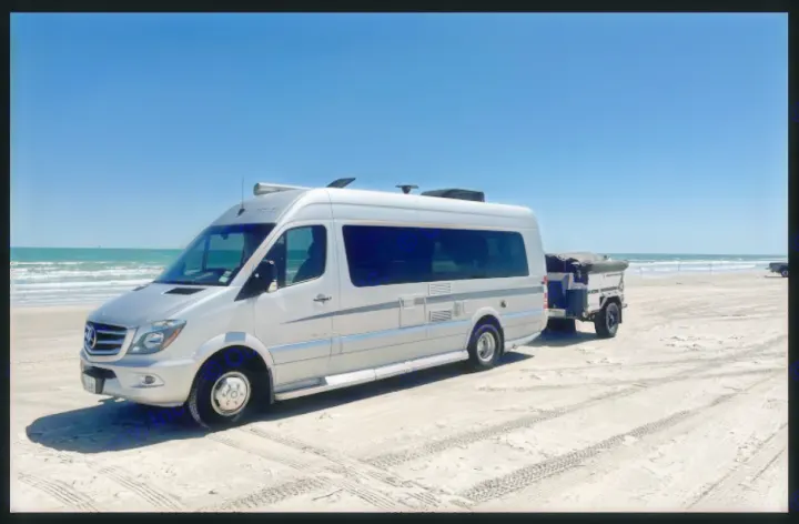 Silver RV rental van & compact trailer on a sunny sandy beach. Ocean waves under clear blue skies, tire tracks visible.. Winnebago Era 2016