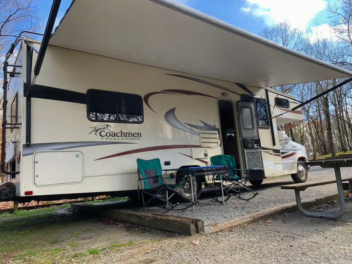 A Class C Coachmen Freelander RV rental with its awning extended at a gravel campsite, featuring camping chairs and a picnic table.. Coachmen Freelander 2016