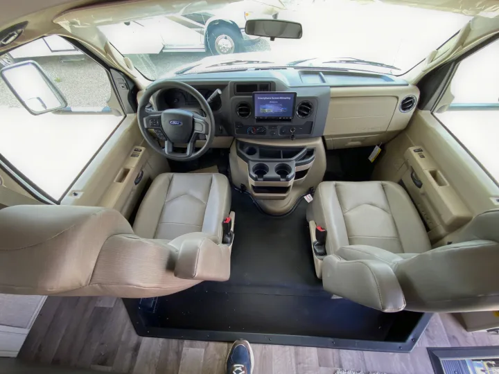 Interior view of an RV rental's front cabin. Features light beige driver and passenger seats, Ford dashboard with screen, and steering wheel.. Jayco Redhawk 2025