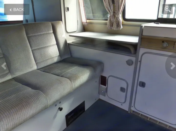 Interior of an RV rental: view of a gray patterned bench seat, fold-out table, and beige kitchen unit with storage and controls.. Volkswagen Westfalia 1990