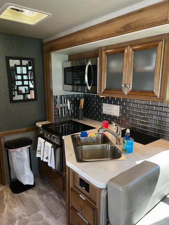 Modern RV rental kitchen with stainless appliances, sink, dark tiled backsplash, wood cabinets, and dinette seating.. Holiday Rambler Admiral 2021