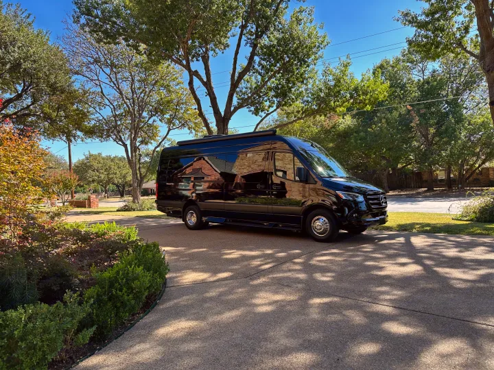 Sleek black Mercedes Sprinter van, an RV rental, parked on a sunny driveway, reflecting homes and trees on its glossy surface.. Ultimate Toys Ultimate RV 2023