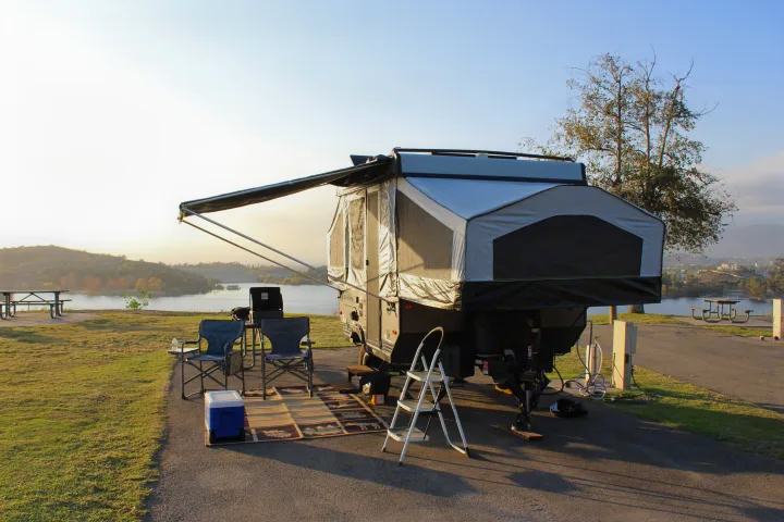 Lakeside camping setup featuring a pop-up RV rental with an extended awning, chairs, and cooler for a relaxing outdoor experience.. Forest River Rockwood ESP 2021