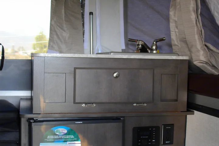 This RV rental's compact kitchen features a sink, faucet, and dark wood cabinets. A Dometic fridge and control panel are seen below.. Forest River Rockwood ESP 2021