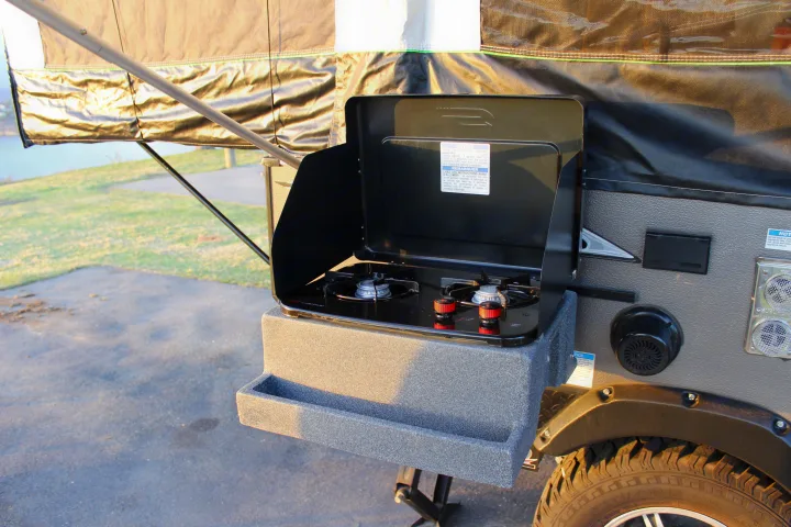 A two-burner outdoor stove attached to an RV rental, ready for camping. Part of a pop-up camper is visible.. Forest River Rockwood ESP 2021