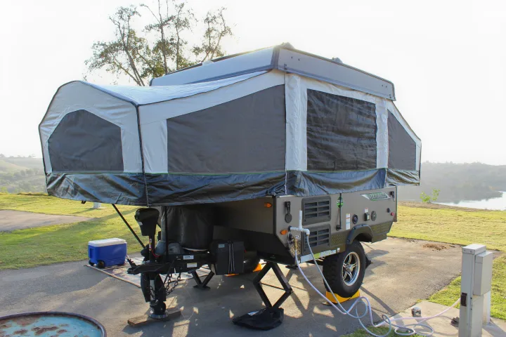 This pop-up RV rental is fully set up at a scenic campsite. Its canvas extensions are deployed, showing hookups and leveling jacks.. Forest River Rockwood ESP 2021