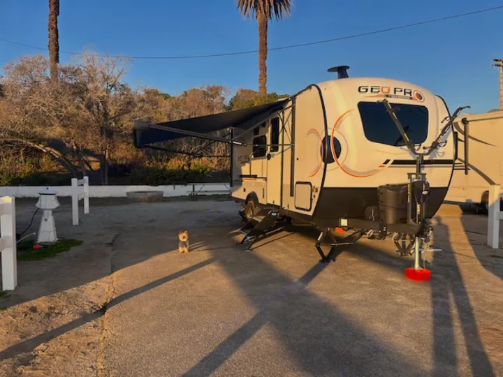 Golden hour at an RV rental campsite. A white GEO PRO travel trailer with an awning is parked, a small dog stands nearby.. Forest River Rockwood Geo Pro 2023
