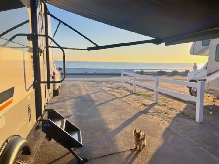 An RV rental by the ocean with steps, awning, and a small dog enjoying the sunset view from a beachside campsite.. Forest River Rockwood Geo Pro 2023