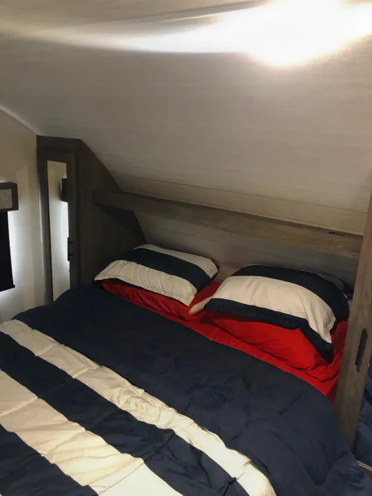 RV rental bed featuring a red, white, and blue striped comforter and overhead storage.. Forest River Wildwood 2020