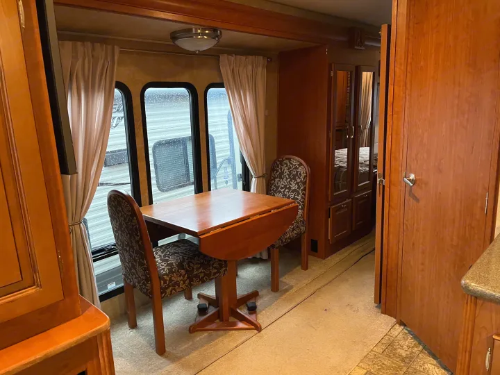 Cozy RV rental dining area with wood table, patterned chairs, and windows showing rain. Bedroom visible beyond.. Forest River Georgetown M373DS 2011