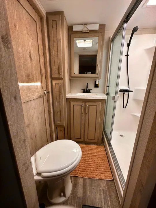 Modern RV rental bathroom: toilet, wood vanity, sink, and glass shower stall. Efficient design for comfortable travel.. Keystone RV Alpine 2022