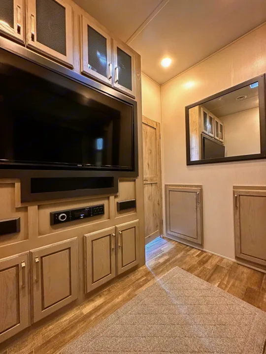 RV rental interior: A large flat-screen TV and audio system are central, surrounded by light wood cabinets. A mirror reflects a kitchen area.. Keystone RV Alpine 2022