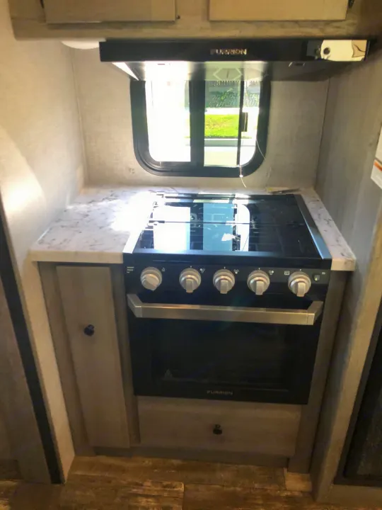 RV rental kitchen shows a Furrion stove/oven, range hood, and marble-look countertop next to a window.. Keystone RV Outback Ultra-Lite 2019