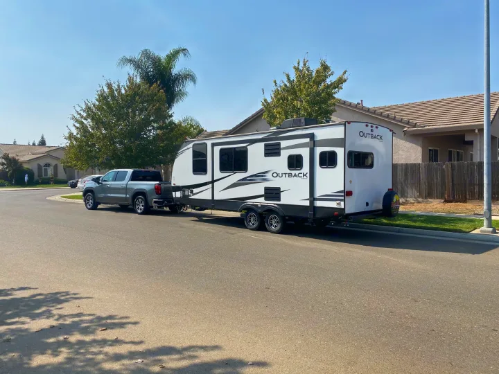 Gray pickup truck towing a white Outback travel trailer on a residential street. Consider this RV for your next rv rental trip!. Keystone RV Outback Ultra-Lite 2019