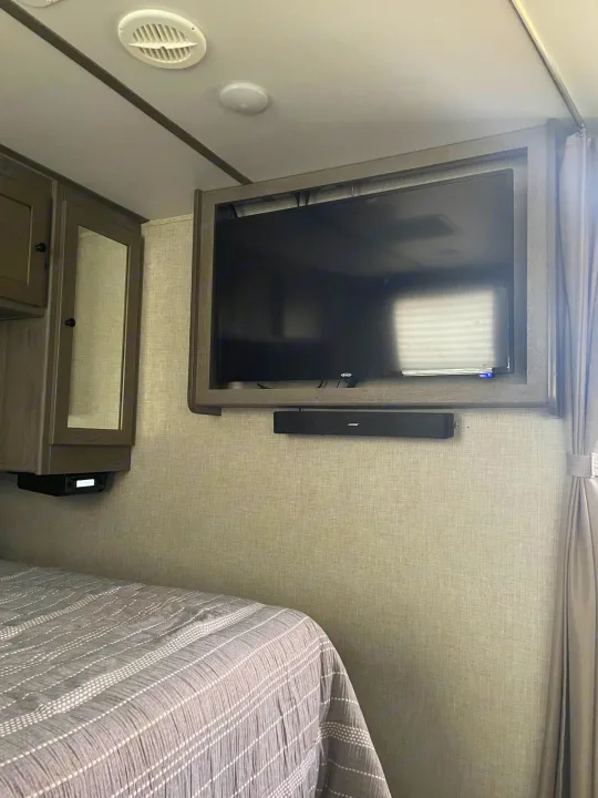 Inside an RV rental: a modern bedroom with a mounted flat-screen TV, Bose soundbar, overhead cabinet, and striped comforter.. Keystone RV Outback Ultra-Lite 2019