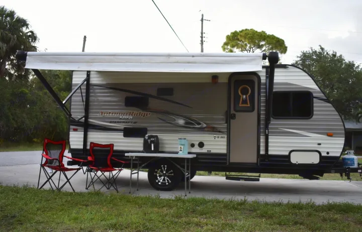 This Autumn Ridge Mini RV rental features an extended awning, two red chairs, and a table with appliances for outdoor enjoyment.. Starcraft Autumn Ridge Mini 2017