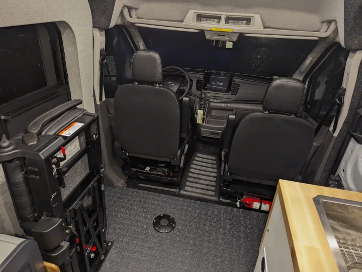 Interior of an RV rental van showing front seats, dashboard, and a deployed wheelchair lift for accessible travel.. Grit Overland GO A/T 2022